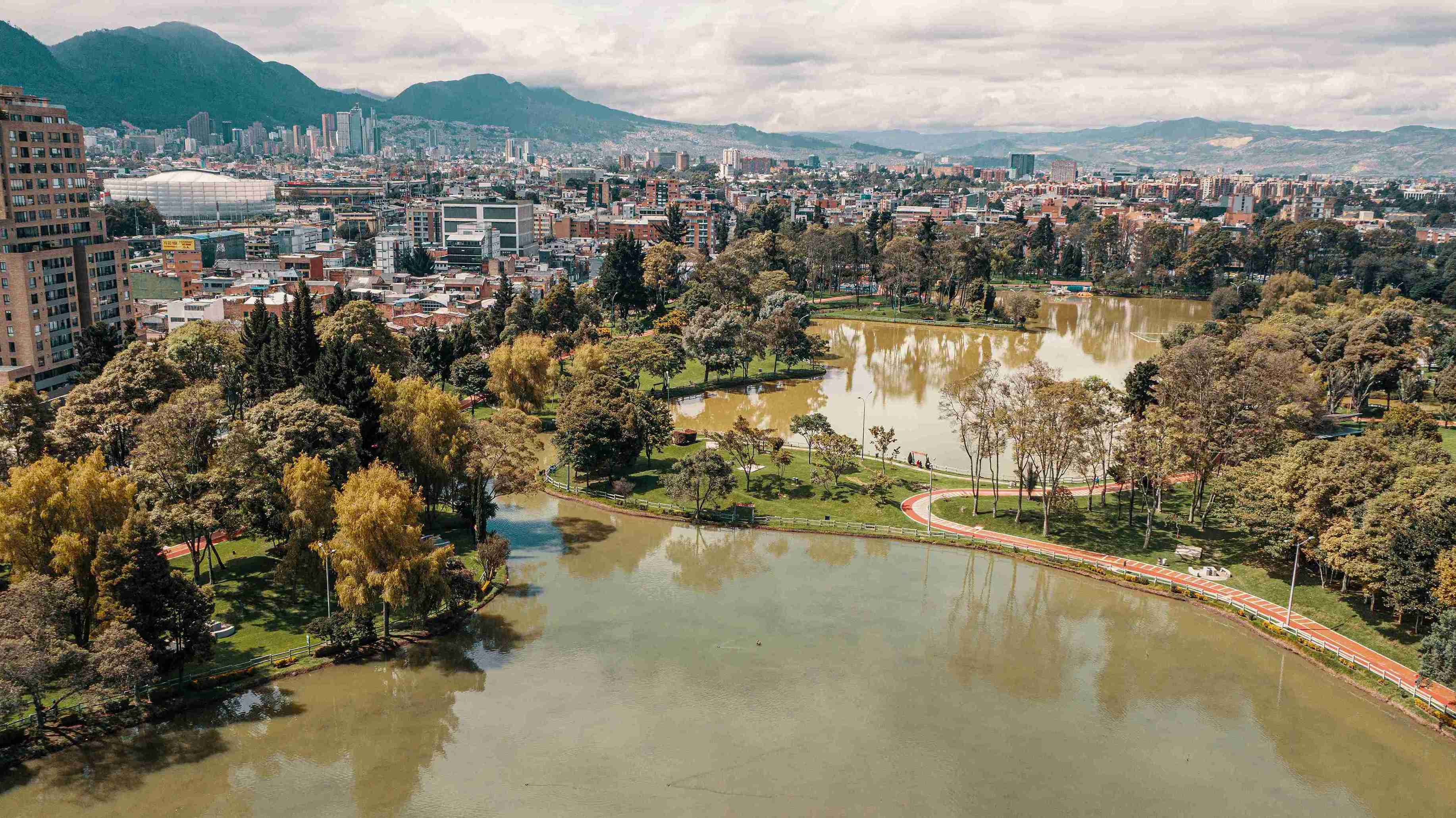 Vista aérea del Parque de los Novios en Bogotá. El lago se mezcla con zonas verdes, senderos rojos para caminar y trotar, y árboles que enmarcan el paisaje con la ciudad y montañas de fondo. Foto: Ricardo Baez - Archivo IDT