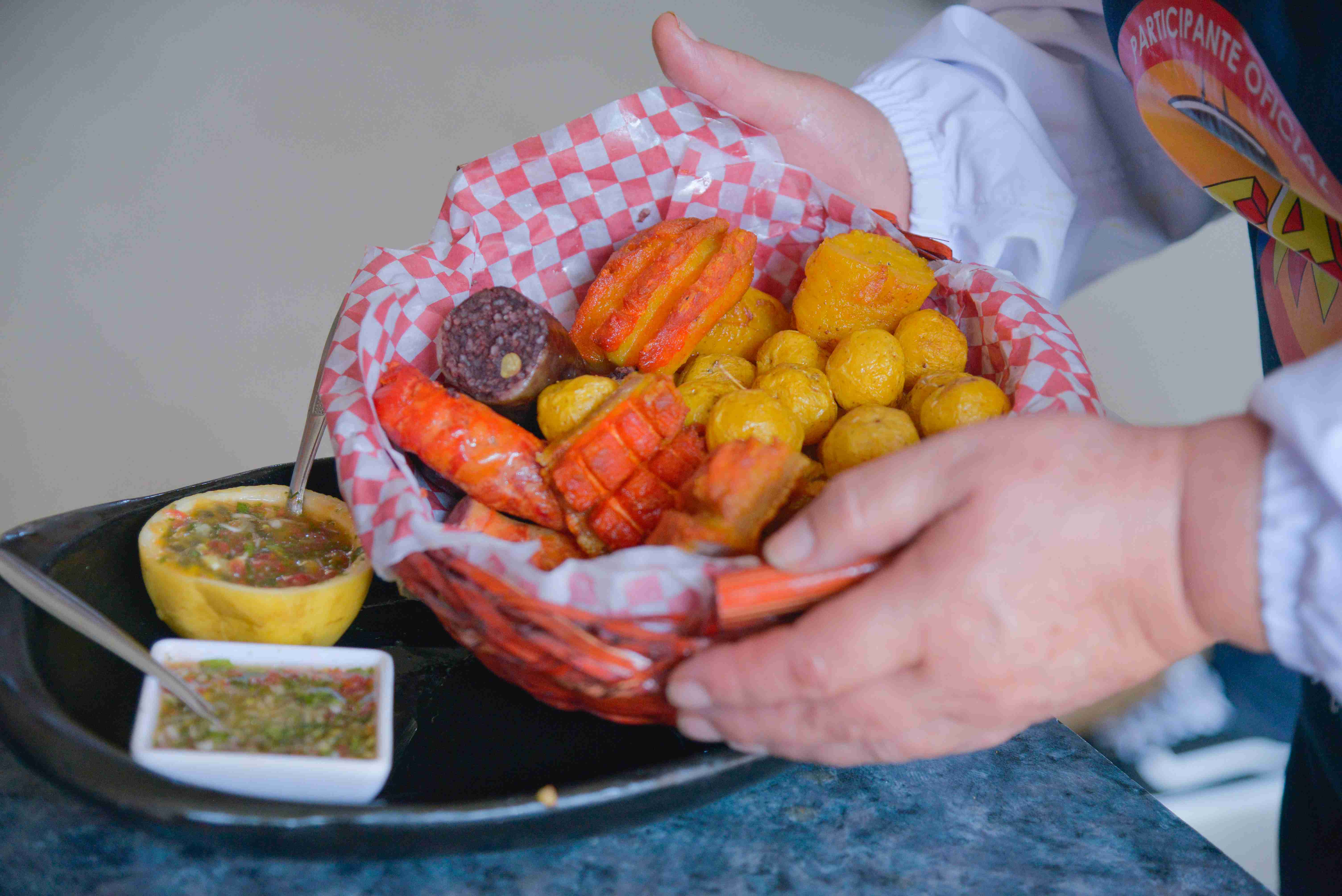 Una canasta colorida llena de fritanga tradicional bogotana. La porción incluye longaniza, chorizo rojo, papa criolla dorada, plátano maduro, morcilla y arepa, acompañada con guacamole y ají casero. Foto: Archivo IDT.